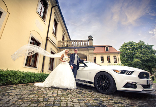 Hochzeitsfotograf Dresden - Das Brautpaar am Mustang vor dem Weingut Proschwitz bei Meißen. Die Braut hat einen wehenden Schleier.
